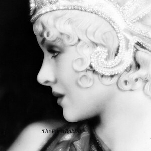 May include: A black and white portrait of a woman wearing a beaded headband and a flowing scarf. The woman is looking to the side and her hair is styled in a short, curly bob.