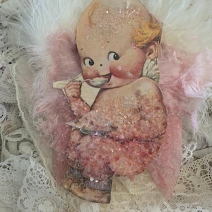 May include: A vintage angel ornament featuring pink feathers and a glittery, pink-toned cherub holding a spoon. The cherub has a large head and a wide smile.