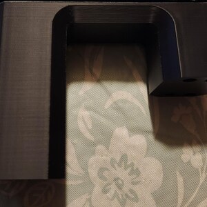 May include: A black plastic 3D printed object with a rectangular shape and a cutout in the middle. The object has a smooth surface and appears to be a part of a larger structure.