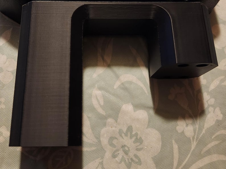 May include: A black plastic 3D printed object with a rectangular shape and a cutout in the middle. The object has a smooth surface and appears to be a part of a larger structure.