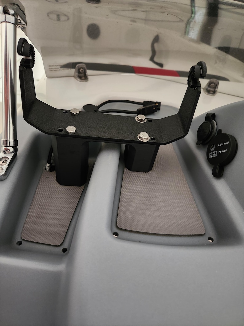 May include: A black metal bracket with two vertical supports and a horizontal bar. The bracket is mounted on a grey surface and has several screws and bolts. The bracket is designed to hold a device, such as a fish finder or GPS unit.