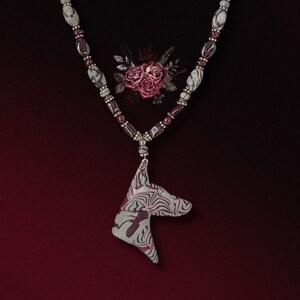May include: A necklace with a white and black marbled pendant shaped like a dog's head. The necklace is made of silver beads and dark red beads.