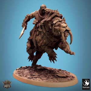May include: A brown, sculpted figurine of an Orc riding a saber-toothed cat. The Orc is holding a sword and wearing armor. The cat is wearing a harness and has a fierce expression.
