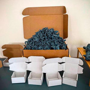 May include: A large brown cardboard box filled with a pile of dark blue miniature figures, accompanied by four empty white cardboard boxes, two empty brown cardboard boxes, and a small wooden table holding additional dark blue figures.