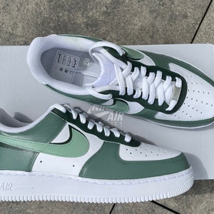 Peut inclure: Une paire de baskets Nike Air Force 1 blanches et vertes. Les baskets ont une semelle blanche, une semelle intermédiaire blanche et une tige verte. Le logo Nike swoosh est noir. Le texte "AIR FORCE 1" est imprimé sur le côté des baskets.
