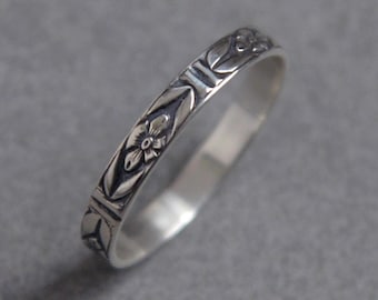 May include: A silver ring with a floral and geometric pattern. The ring features a repeating design of flowers and angular shapes around the band. The ring is set against a gray background, highlighting the intricate details of the design.