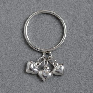 May include: A silver ring featuring three heart-shaped charms. The ring and charms are made of polished metal. The hearts are small and rounded, with a smooth surface. The ring is set against a grey background.