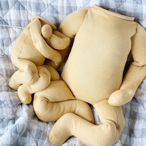 May include: A doll body and limbs in a light yellow fabric, lying on a gray and white checkered quilt. The doll parts include a torso, arms, and legs, all unstuffed. The text "Reggiedolls" is visible in the lower left corner.