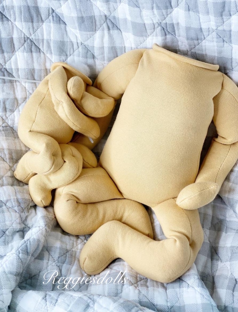 May include: A doll body and limbs in a light yellow fabric, lying on a gray and white checkered quilt. The doll parts include a torso, arms, and legs, all unstuffed. The text "Reggiedolls" is visible in the lower left corner.