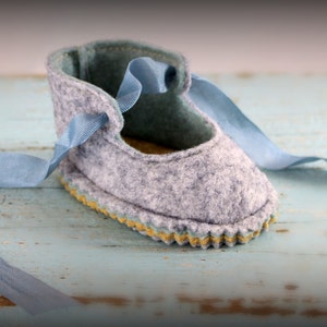 Puede incluir: Un par de botines de bebé de fieltro gris con cordones de cinta azul. Los botines tienen un detalle de costura amarilla alrededor del borde.