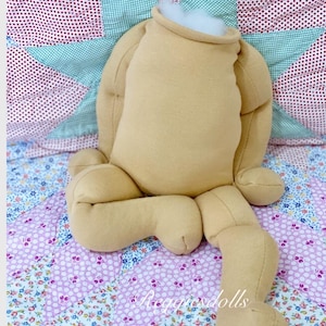 May include: A tan doll body with arms and legs, stuffed with white filling at the neck. The doll is positioned on a floral patterned quilt with a star-shaped design in the background. The doll is unfinished.