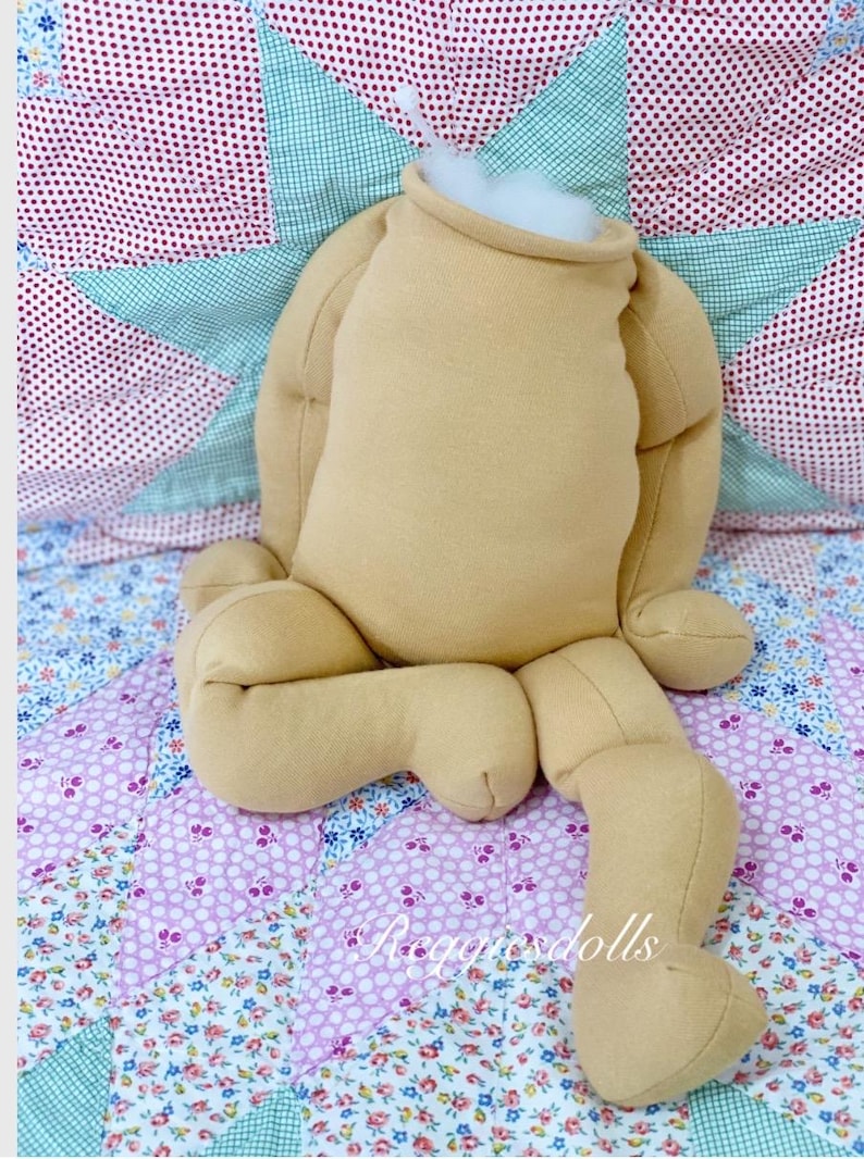 May include: A tan doll body with arms and legs, stuffed with white filling at the neck. The doll is positioned on a floral patterned quilt with a star-shaped design in the background. The doll is unfinished.