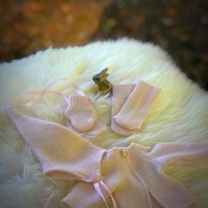 May include: A pink knitted baby cardigan with a pearl button closure and matching pink knitted mittens. The cardigan has a pink ribbon tied around the waist. The cardigan is laid out on a white fluffy surface.