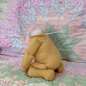 May include: A tan-colored doll body with attached limbs, stuffed with white material at the head. The doll is positioned on a patchwork quilt with floral and star patterns. The text "Reggiesdolls" is visible.