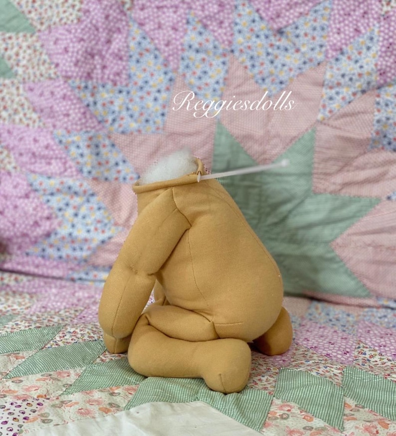 May include: A tan-colored doll body with attached limbs, stuffed with white material at the head. The doll is positioned on a patchwork quilt with floral and star patterns. The text "Reggiesdolls" is visible.