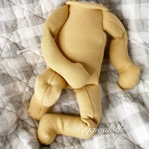 May include: A yellow doll body with attached arms and legs, made of soft fabric. The doll body is lying on a gray and white checkered quilt. The text "Reggiesdolls" is printed on the leg.