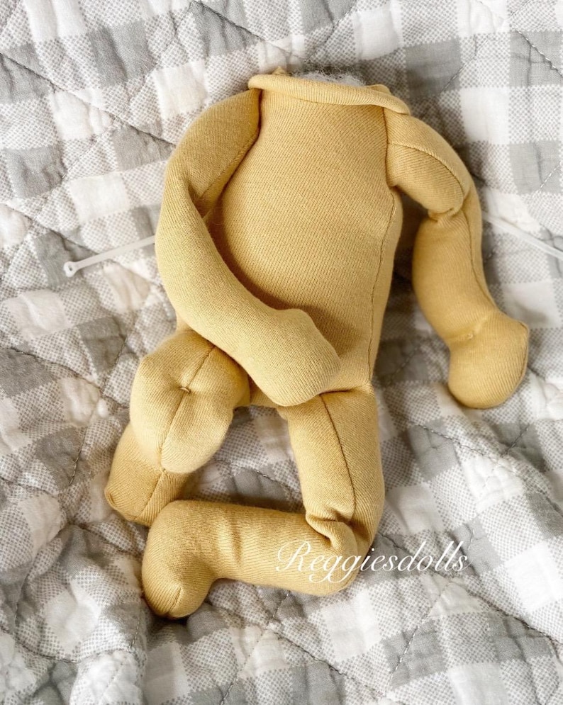 May include: A yellow doll body with attached arms and legs, made of soft fabric. The doll body is lying on a gray and white checkered quilt. The text "Reggiesdolls" is printed on the leg.
