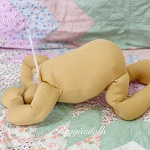 May include: A tan-colored doll body with arms and legs, lying on a floral patterned quilt. The doll's limbs are bent, and a white plastic tie is attached to the doll. The text "reggiedolls" is visible in the bottom right corner.