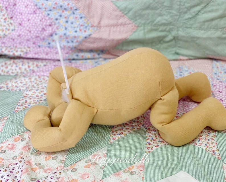 May include: A tan-colored doll body with arms and legs, lying on a floral patterned quilt. The doll's limbs are bent, and a white plastic tie is attached to the doll. The text "reggiedolls" is visible in the bottom right corner.