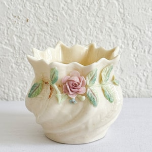 May include: A small, cream-colored ceramic vase with a ruffled rim and a floral design. The vase features a pink rose with green leaves and a swirl pattern.
