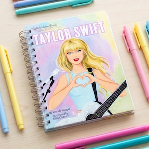 May include: A spiral-bound notebook with a cover featuring a portrait of Taylor Swift, surrounded by colorful pens in shades of pink, blue, yellow, and green. The notebook has the text "Taylor Swift" on the cover.