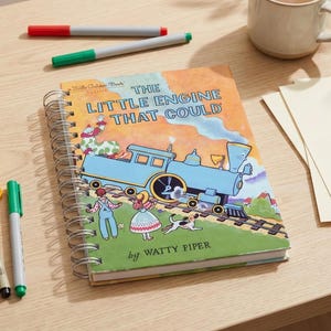 May include: A still-life image featuring a copy of "The Little Engine That Could" book, a mug of coffee, and colorful markers. The book has a blue train illustration. Markers are red, blue, yellow, and green. A ruler and blank papers are also present.
