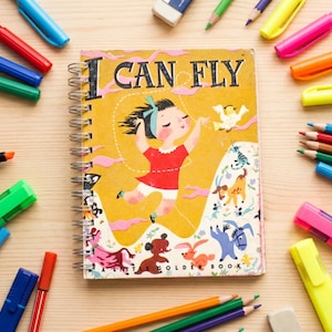 Puede incluir: Un libro infantil con espiral titulado "I Can Fly" en una portada amarilla, rodeado de bolígrafos, lápices y rotuladores de colores. La ilustración del libro muestra a una niña con un vestido rojo.