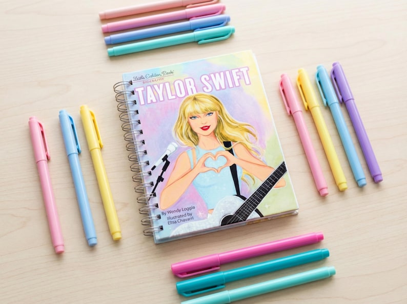 May include: A spiral-bound notebook with a cover featuring a portrait of Taylor Swift, surrounded by colorful pens in shades of pink, blue, yellow, and green. The notebook has the text "Taylor Swift" on the cover.