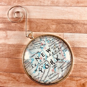 May include: A gold-colored ornament featuring a map of New York State with the words "NEW YORK" in large letters. The ornament is round and has a clear glass front.