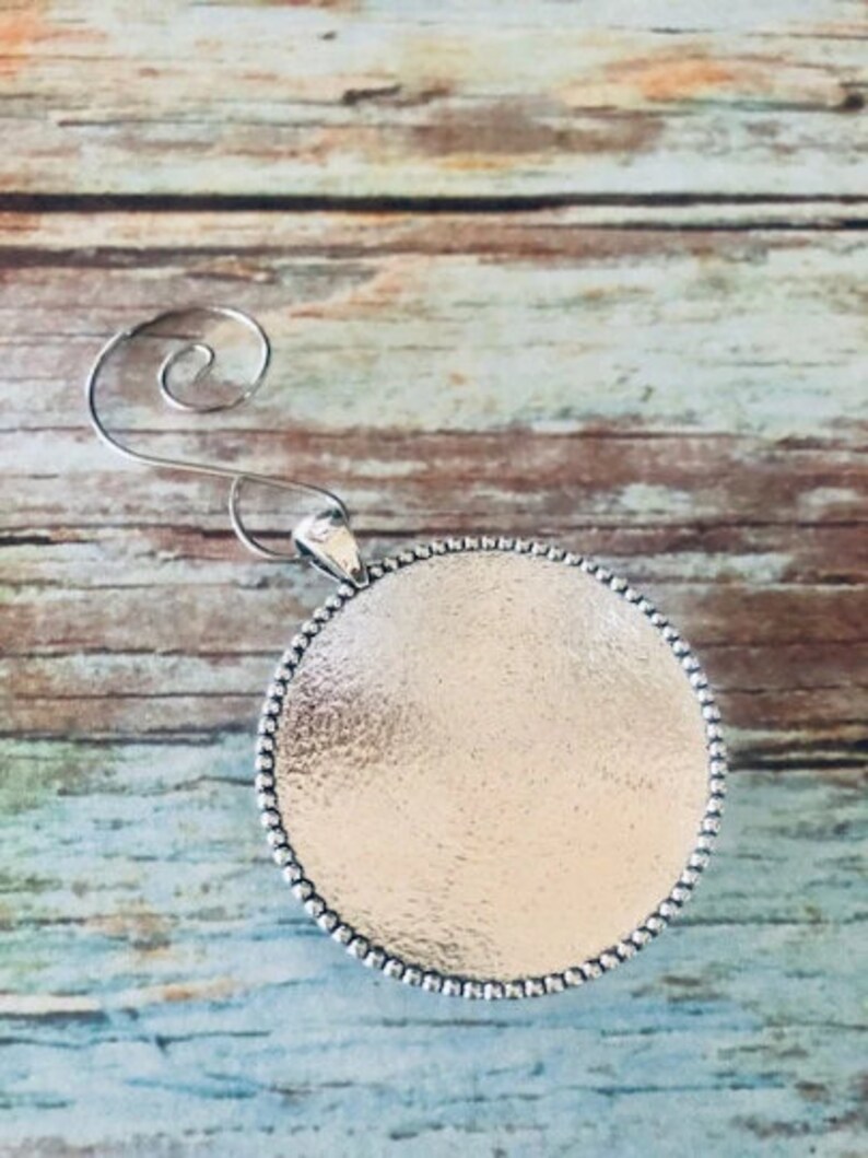 May include: Silver ornament with a round, textured surface and a beaded edge. The ornament has a wire hanger for hanging.
