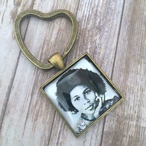 May include: A square pendant with a black and white portrait of a person, set in a bronze-colored frame. The pendant is attached to a heart-shaped key ring, also in a bronze finish. The image is on a light wood surface.