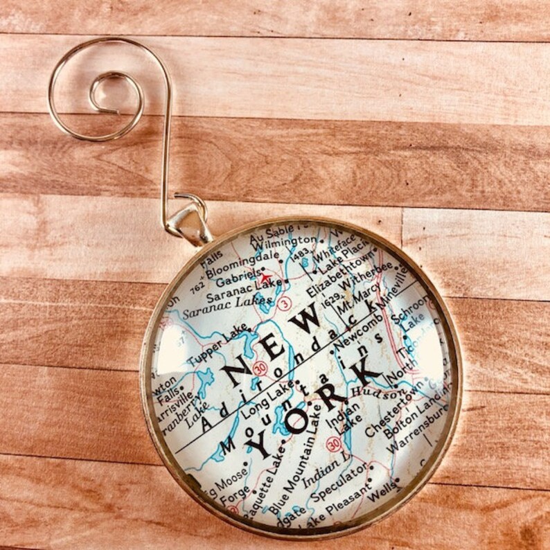 May include: A gold-colored ornament featuring a map of New York State with the words "NEW YORK" in large letters. The ornament is round and has a clear glass front.