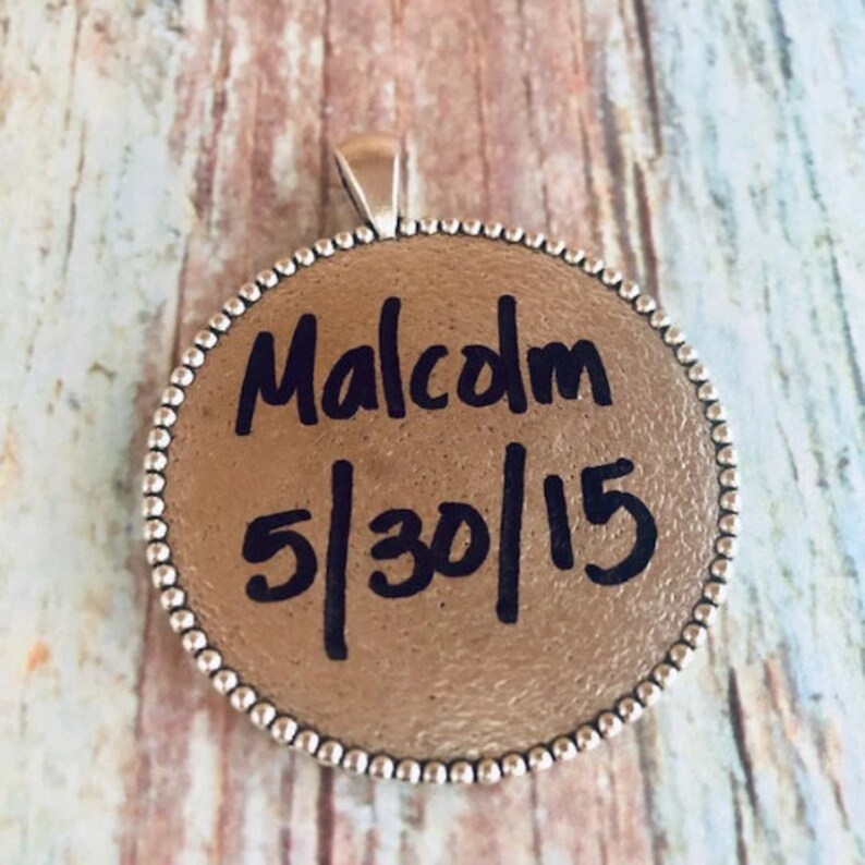 May include: A silver round pendant with a beaded edge. The pendant has black ink writing that says "Malcolm 5/30/15".