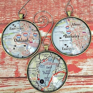 May include: Three round glass ornaments with gold trim, each featuring a map of a different California city. The first ornament shows a map of Oakland, the second shows a map of Lake Tahoe, and the third shows a map of Berkeley.