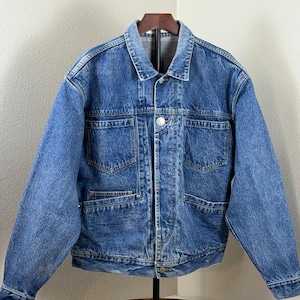 May include: A medium-wash blue denim jacket with a classic collar, button closure, and multiple pockets. The jacket is displayed on a wooden hanger and has a vintage aesthetic. The jacket has two chest pockets and two lower pockets.