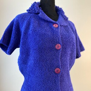 May include: A blue short-sleeved cardigan with a fuzzy texture and a collar. The cardigan has five purple buttons down the front.