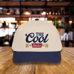 May include: A baseball cap with a cream-colored crown and a navy blue brim. The cap features the text "The Cool Uncle" in navy blue script, with "Est. 2026" and a decorative emblem below. The cap is sitting on a wooden surface.