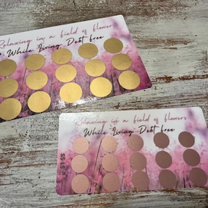 May include: Two rectangular cards with a floral design and gold scratch-off circles. The text "Relaxing in a field of flowers While Living Debt free" is printed on the cards. The cards have a pink and white background.