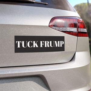 May include: Black rectangular bumper sticker with the words "TUCK FRUMP" in white, sans-serif font. The sticker is affixed to the rear of a silver car, near the taillight.