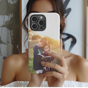 May include: White phone case featuring a photograph of two young girls reading a book outdoors. The image shows the girls in a field with a warm, golden sunset in the background. The phone case has a glossy finish.