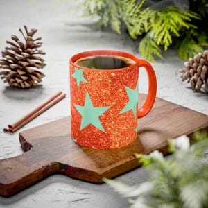 May include: A red ceramic mug with a glittery texture and large, light blue star shapes. The mug has a red handle and is filled with dark liquid, possibly coffee or tea. The mug is on a wooden cutting board.