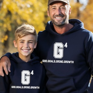 May include: Two people wearing navy blue hooded sweatshirts with a white "G4" logo and the text "GOD.GOALS.GRIND.GROWTH" on the front. The people are standing in a forest with fall foliage.