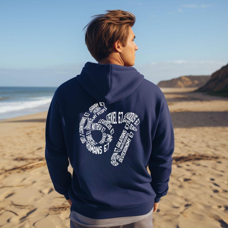 May include: Navy blue hoodie with white text on the back. The text includes the number 67 and various biblical references. The hoodie has a hood and a front pocket. The person in the image is standing on a sandy beach.