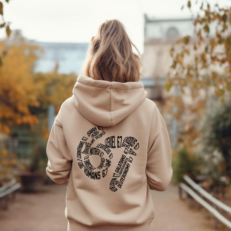 May include: A beige hoodie with a hood, featuring a black graphic on the back. The graphic is a stylized number 67 composed of various biblical verses. The person is facing away from the camera, walking on a path with trees in the background.