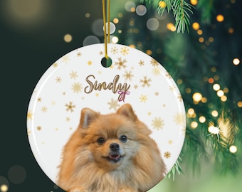 May include: A white ceramic Christmas ornament with a golden snowflake pattern and the name "Sindy" written in cursive. A fluffy, light brown Pomeranian dog is featured in the center of the ornament.