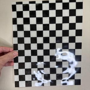 May include: A clear, rectangular sheet with a black and white checkerboard pattern. The pattern consists of evenly spaced squares, creating a classic design. The sheet is held against a neutral background.