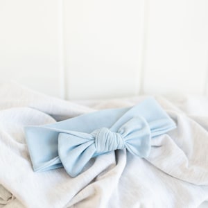 May include: A light blue baby headband with a large bow. The headband is made of soft fabric and is laying on a white and gray textured blanket. The bow is knotted in the center.