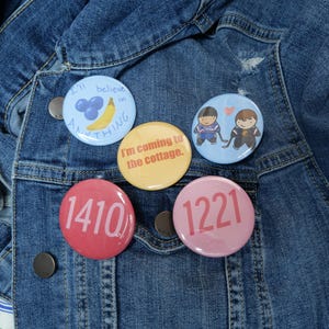 Könnte beinhalten: Fünf runde Ansteckbuttons mit verschiedenen Designs und Texten werden auf einer Jeansjacke gezeigt. Die Buttons sind hellblau, gelb, rosa und rot. Ein Button sagt "I'll believe in ANYTHING" mit einer Banane und Blaubeeren. Andere Buttons haben Zahlen und Cartoonfiguren.