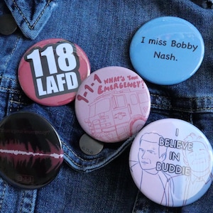 Peut inclure: Plusieurs badges ronds avec des motifs et des textes variés, dont "118 LAFD", "I miss Bobby Nash" et "I BELIEVE IN BUDDIE". Les badges sont roses, bleus et noirs, posés sur une veste en jean.