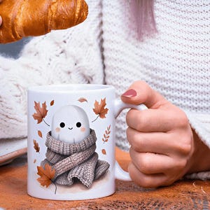 May include: White ceramic mug with a cute ghost character in a scarf and autumn leaves. The mug is held by a person. A croissant is held in the other hand. The mug is on a wooden surface. The mug has a handle.